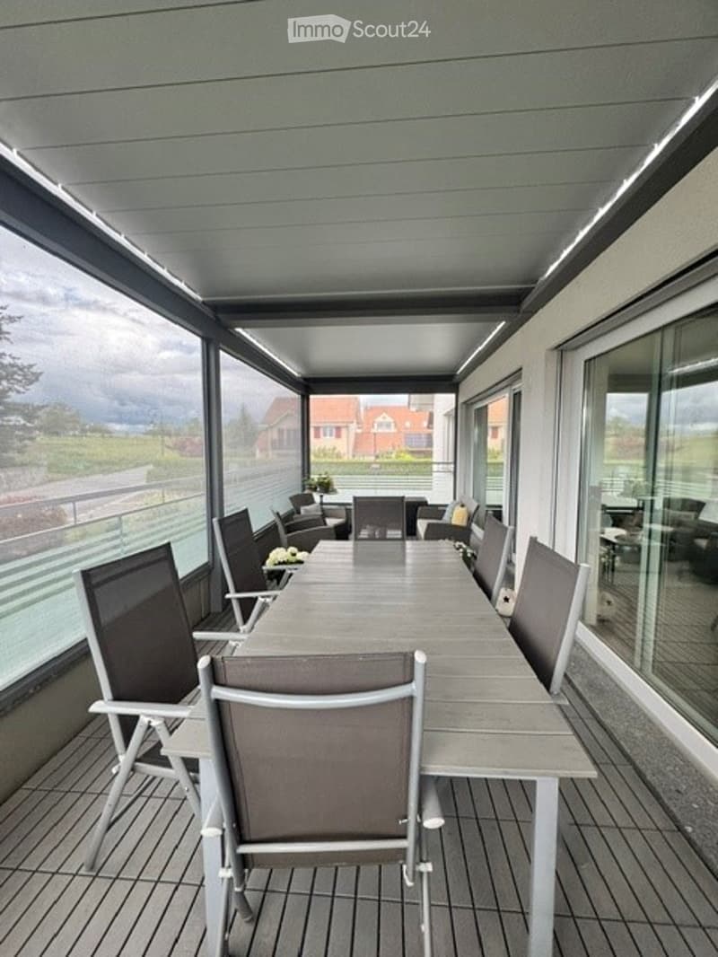 Covered balcony with a long wooden table and several chairs, overlooking a residential area with houses and greenery.
