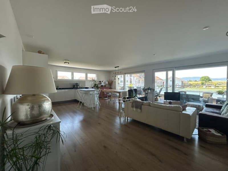 Open-plan living and dining area with wooden flooring, large windows offering a scenic view, a cream-colored sofa, a coffee table, dining chairs, a lamp, and a screen.