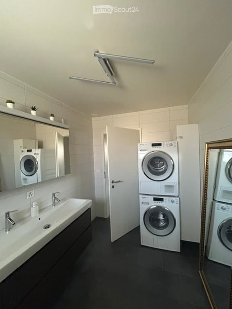 Modern bathroom with a white sink, mirror, and a stacked washing machine and dryer unit. The space features white tiles and a sleek design.