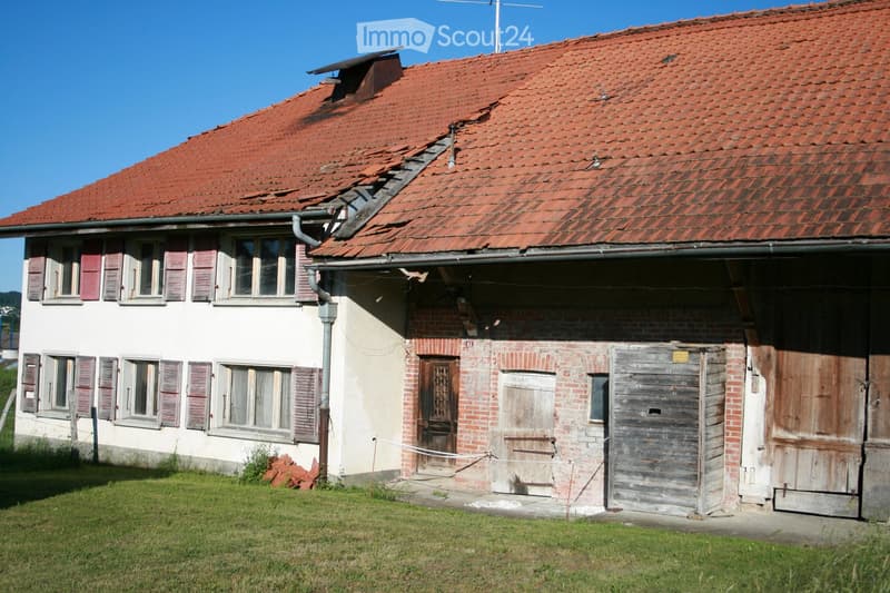 Two-story house with red-tiled roof, white exterior walls, multiple windows with shutters, and a brick garage section with wooden doors.