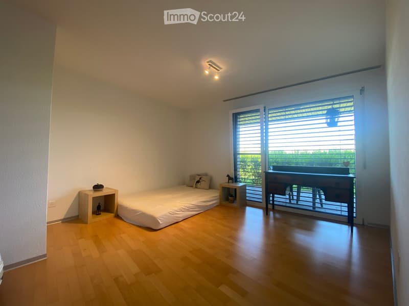 Minimalist bedroom with a single bed, nightstand, small table, and a view through large windows with blinds.