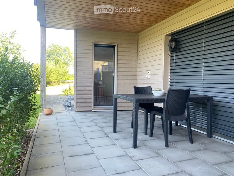 Outdoor patio with a wooden ceiling, tiled floor, and a black wicker dining set including a table and chairs. A sliding glass door leads to the interior, and there are potted plants and greenery nearby.