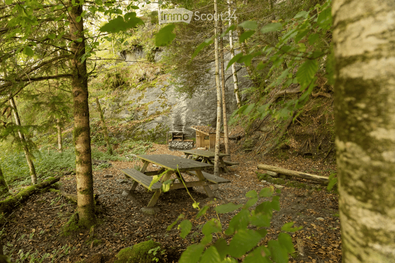 A serene outdoor area with wooden picnic tables, a small wooden shelter, and a stone fireplace nestled among trees and rocky terrain.