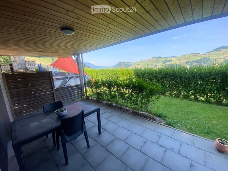 Balcony with a table, chairs, and potted plants, overlooking a scenic landscape with hills and mountains.