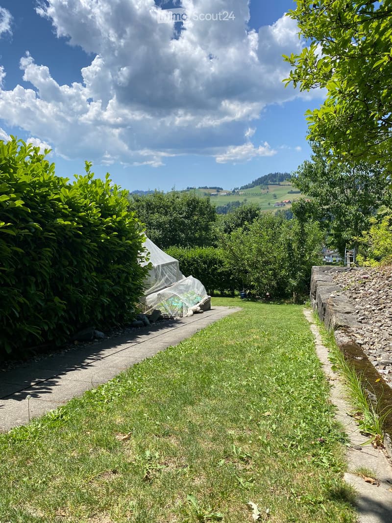 A well-maintained garden with a concrete pathway, lush green grass, and a stone wall on one side. The background features a scenic view of rolling hills and trees under a partly cloudy sky.