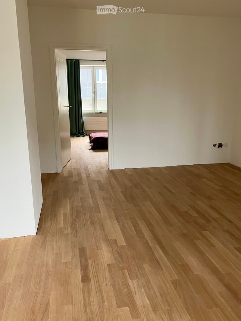 Empty hallway with light wooden flooring and white walls, leading to a bedroom with a bed and green curtains.