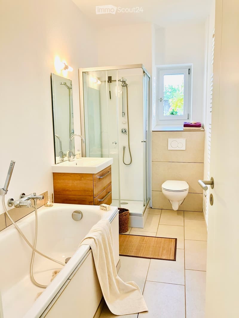 Modern bathroom with a bathtub, toilet, sink, shower, and wooden cabinet. Features a large mirror, towel on the edge of the tub, and a window with a view of greenery.