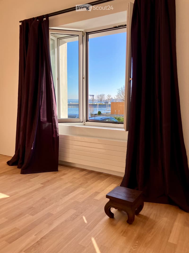 Empty room with wooden flooring, large window with dark curtains, and a small wooden stool near the window. The view shows a waterfront with a clear blue sky.