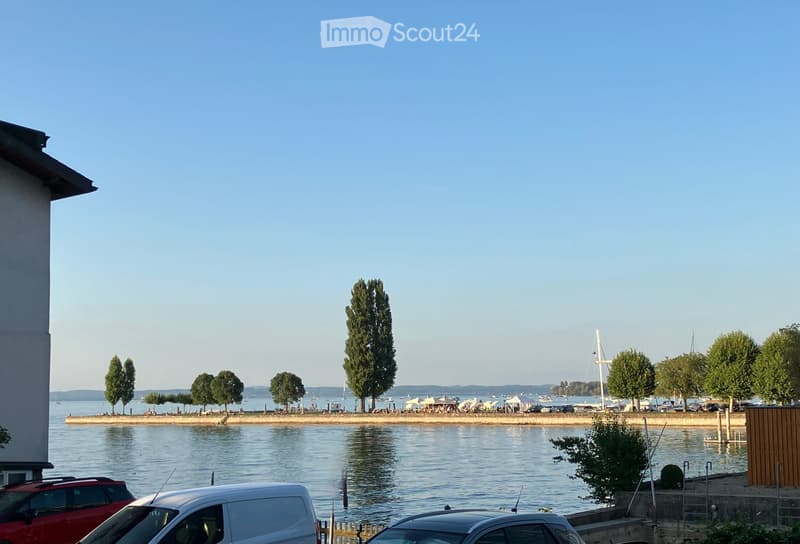 A waterfront view with calm water, trees, boats, and parked cars along the shore.
