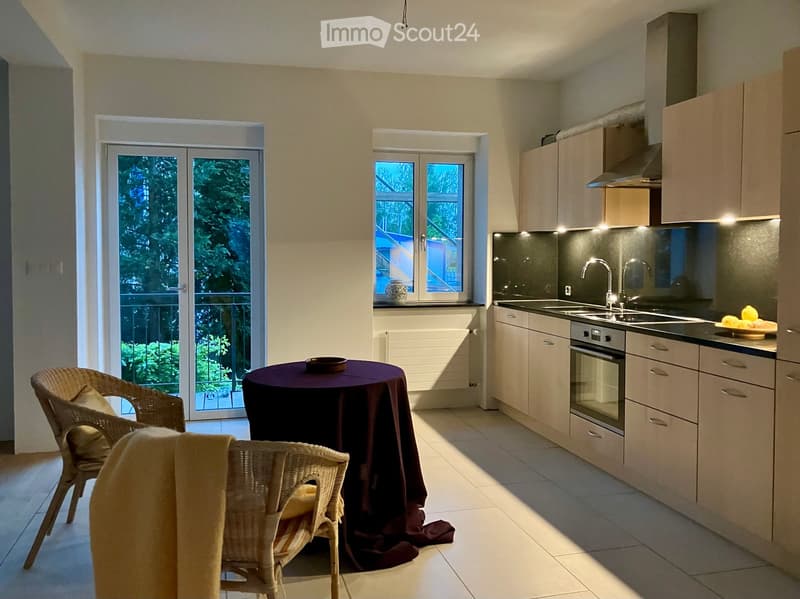 Modern kitchen with white cabinets, stainless steel appliances, and a dining area with a round table and wicker chairs. The space features large windows and a sliding glass door leading to a balcony.