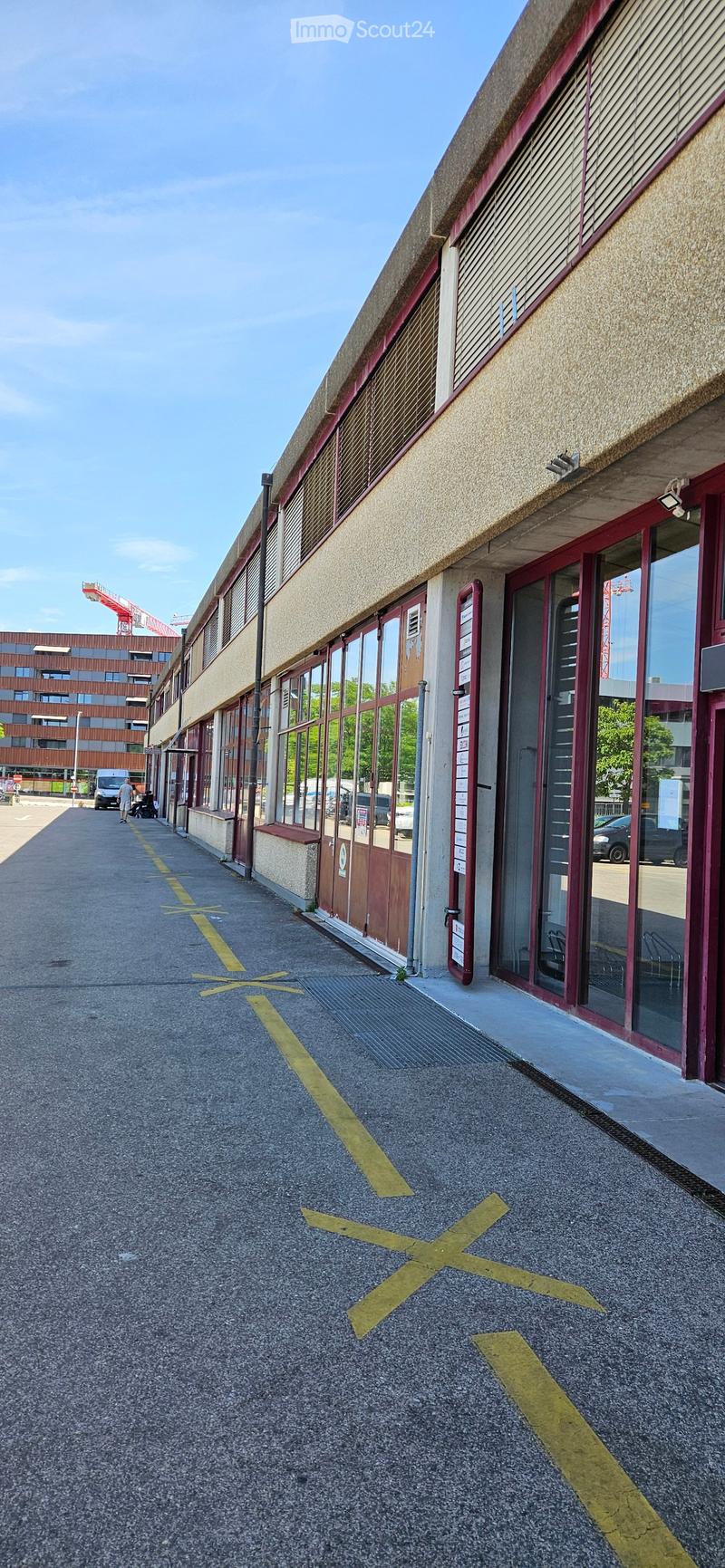 Modern building entrance with large glass doors, red-framed windows, and a paved area with yellow directional markings.