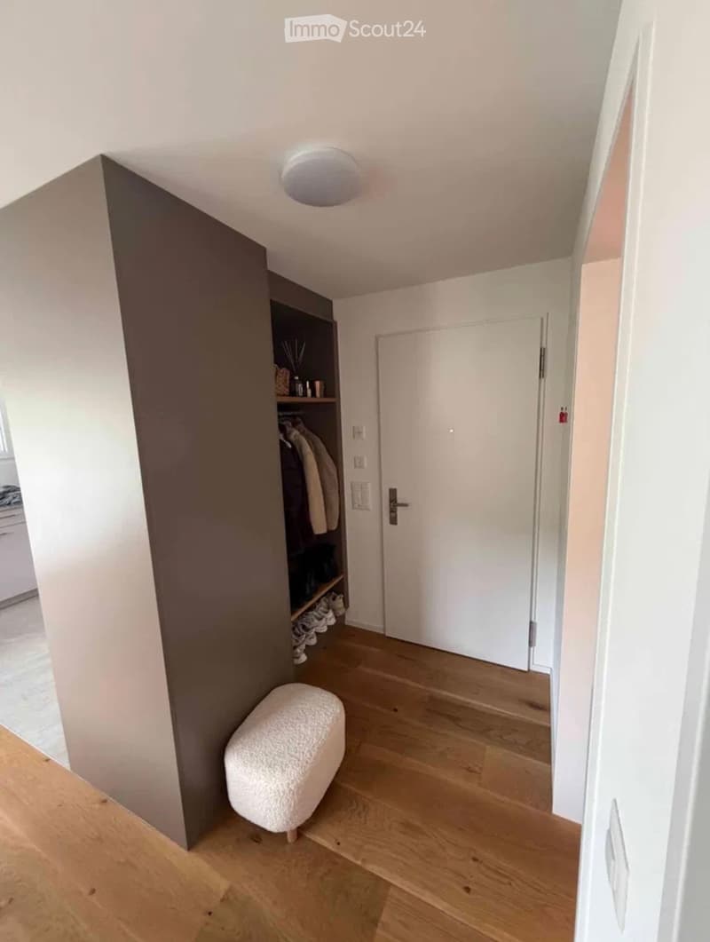 A modern hallway with light wooden flooring, white walls, a built-in closet with clothes and shoes, a small white bench, and a closed door leading to another room.