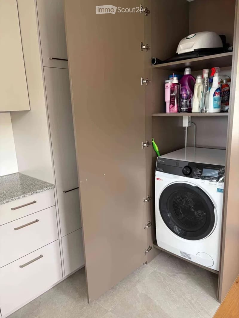A compact laundry area with a white washing machine, cleaning supplies on shelves, and light-colored cabinets.