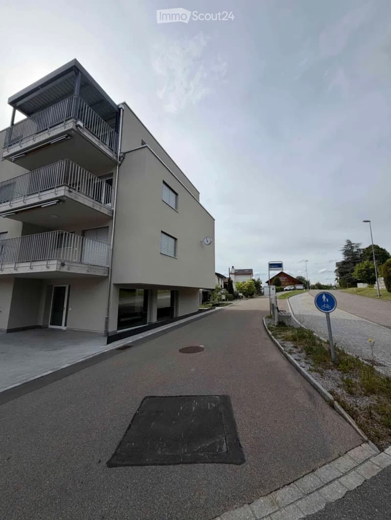 Modern two-story building with balconies, grey facade, and large windows. The entrance has a glass door, and the building is situated on a paved street with a bicycle sign nearby.