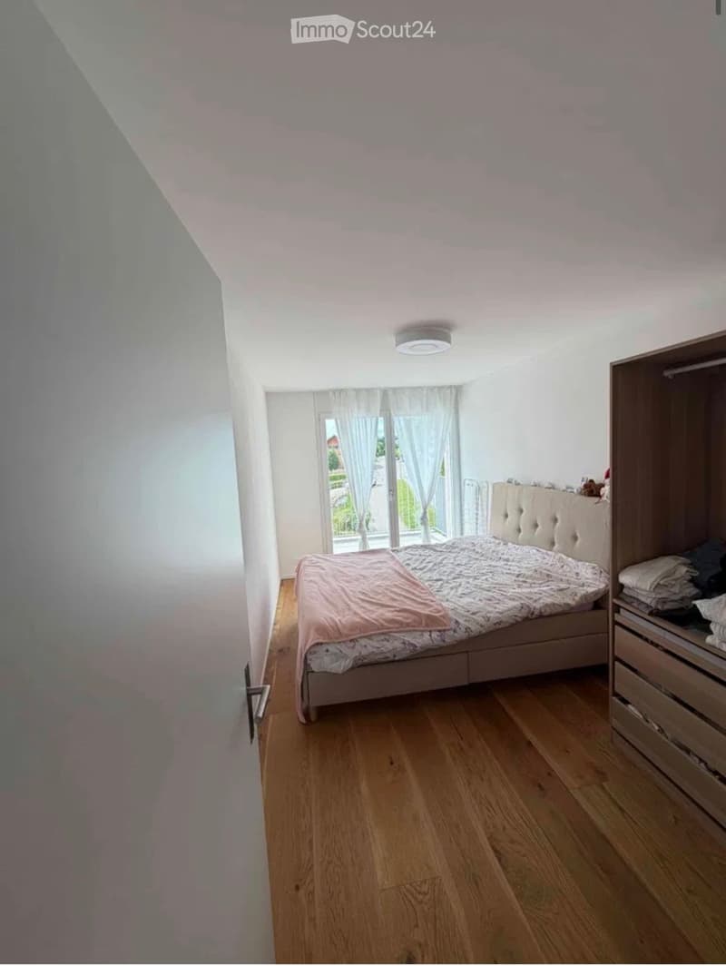 Bright bedroom with wooden flooring, a large bed with a tufted headboard, a built-in wardrobe, and large windows with white curtains.