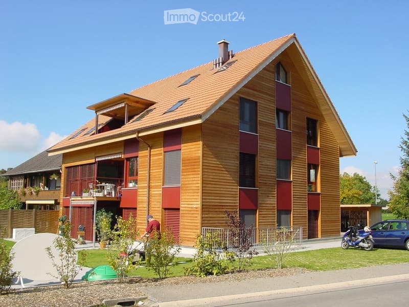 Two-story house with wooden facade, red accents, and a tiled roof. Features a balcony, garage, and parking area.