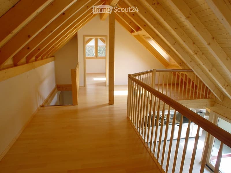 Bright, wooden-floored hallway with exposed wooden beams and a wooden railing leading to an open area with large windows.