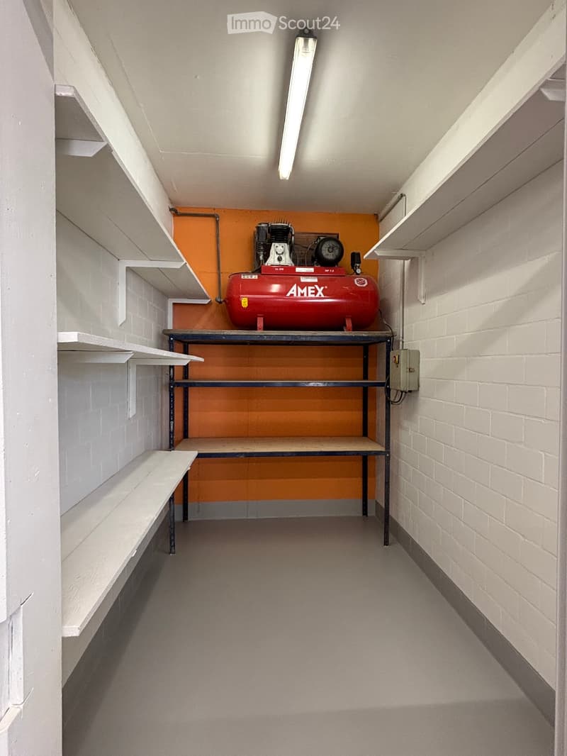 A small, well-lit storage room with white brick walls, a grey concrete floor, and built-in white shelves. A red air compressor sits on a metal table in the center.
