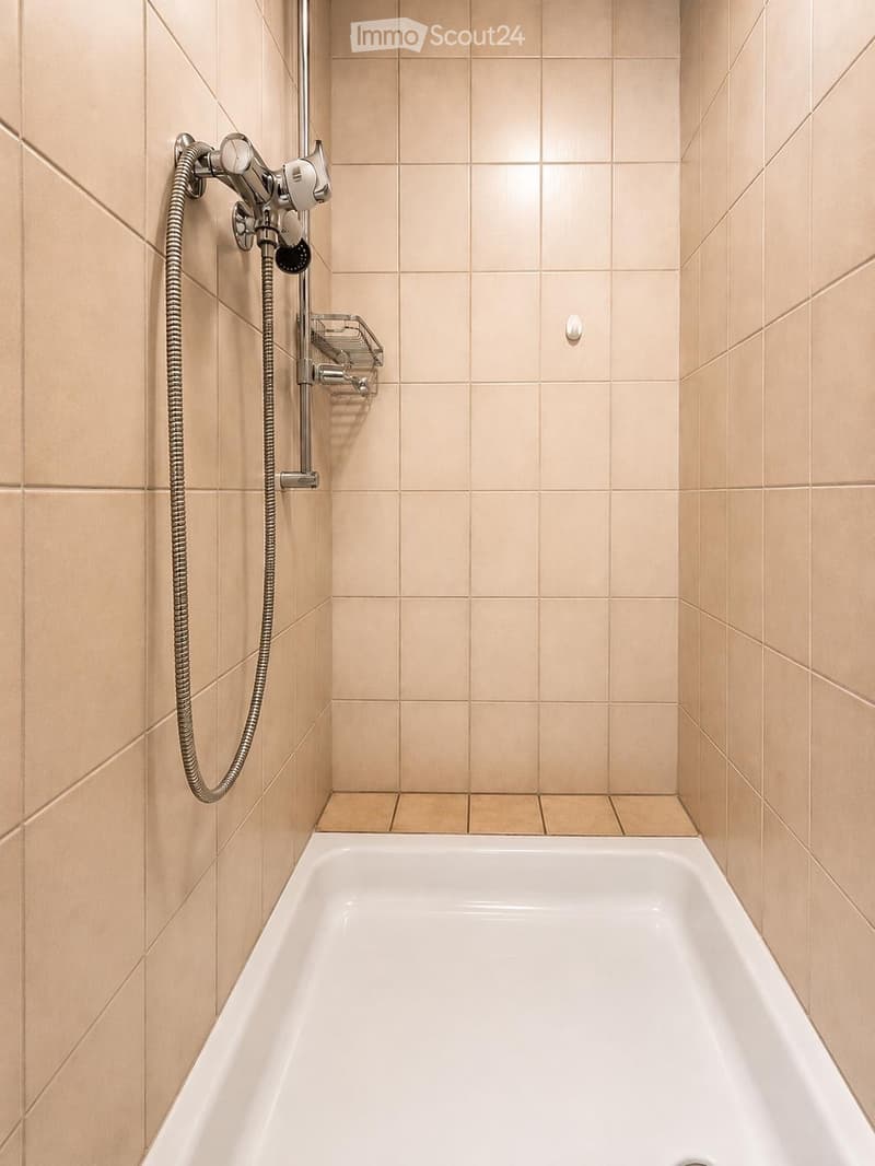 Modern bathroom with a white bathtub, beige tiled walls, and a handheld showerhead with a hose and shelf.