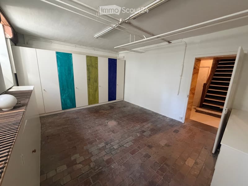 Empty room with white walls, exposed brick floor, white cabinetry, and a staircase leading to another level.
