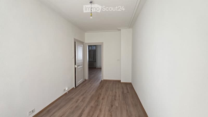 Empty room with wooden flooring, white walls, and a single light fixture hanging from the ceiling. There is a doorway leading to another room.