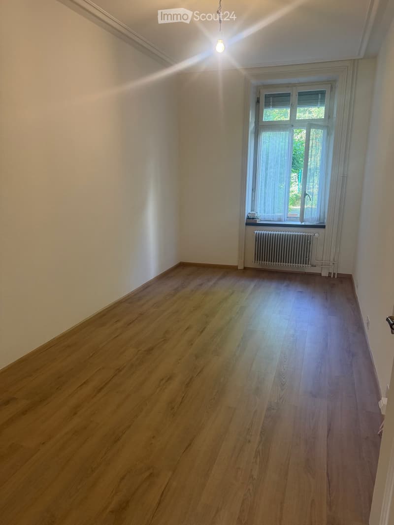 Empty room with wooden flooring, white walls, a window with white frame and curtains, a radiator, and a hanging light fixture.