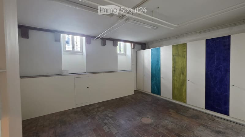 Empty room with white walls, exposed ceiling pipes, and a wooden floor. Features include a small window, a cabinet, and colorful wall panels.