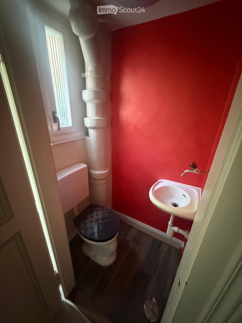 Small bathroom with red walls, a toilet with a dark seat cover, a white sink with a chrome faucet, and a window with white blinds.