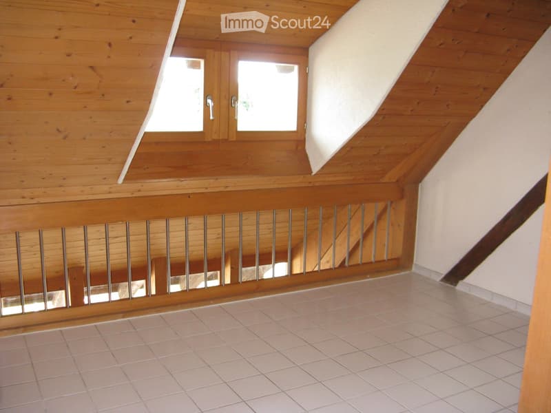 Empty room with wooden walls, slanted wooden ceiling, tiled floor, and a metal railing leading to another area.