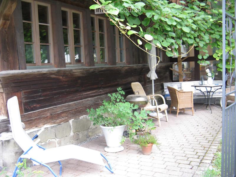 Outdoor patio area with wooden house backdrop, featuring a lounge chair, wicker chairs, a small table, potted plants, and a grill.