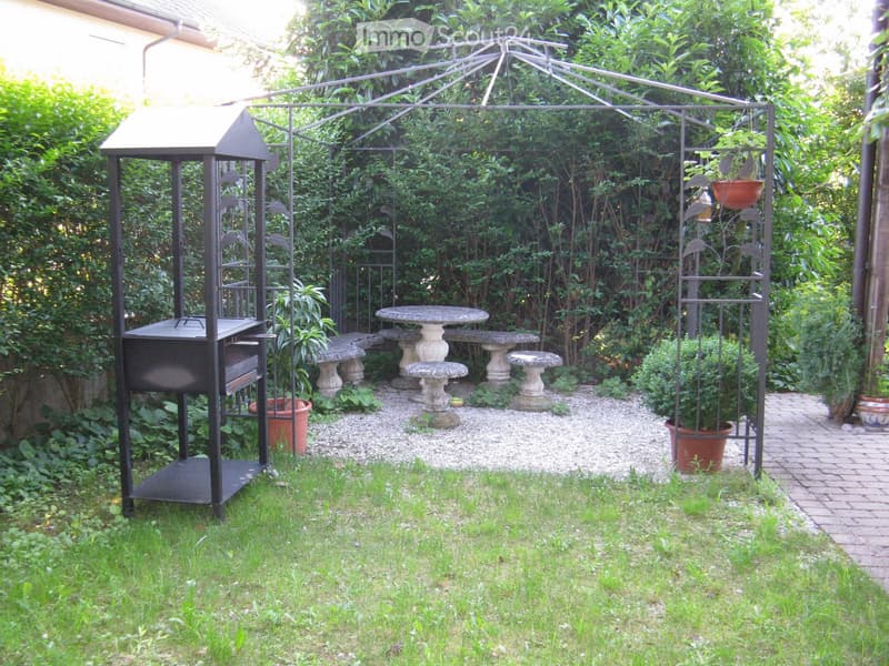 Garden with a metal gazebo, stone seating area, barbecue, potted plants, and gravel pathways.