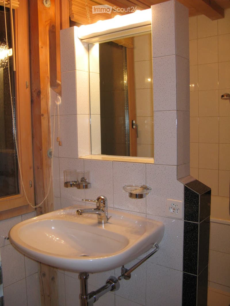 Modern bathroom with a white sink, chrome faucet, mirror with built-in lighting, toothbrush holder, and soap dish. The walls are tiled with light-colored tiles and a dark accent strip.