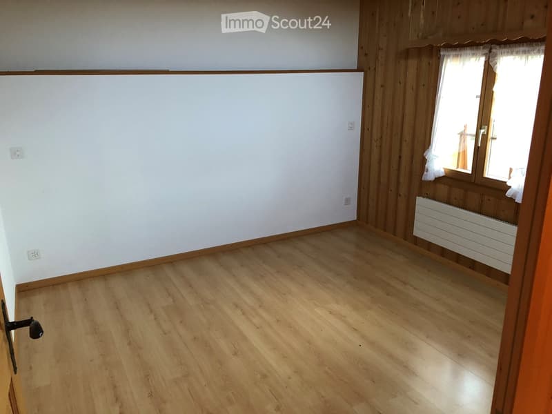 Empty room with wooden flooring, white walls, and large windows with white curtains. Radiator visible on the wall.