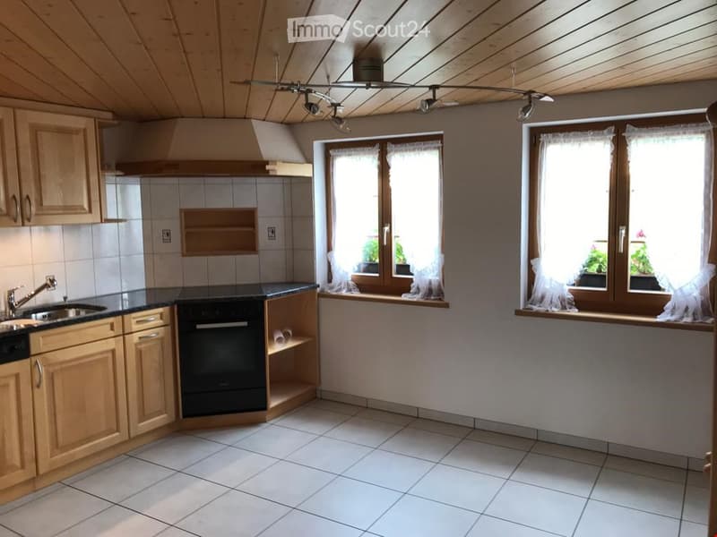 Bright kitchen with wooden cabinets, tiled floor, and white walls. Features a sink, oven, and large windows with sheer curtains.