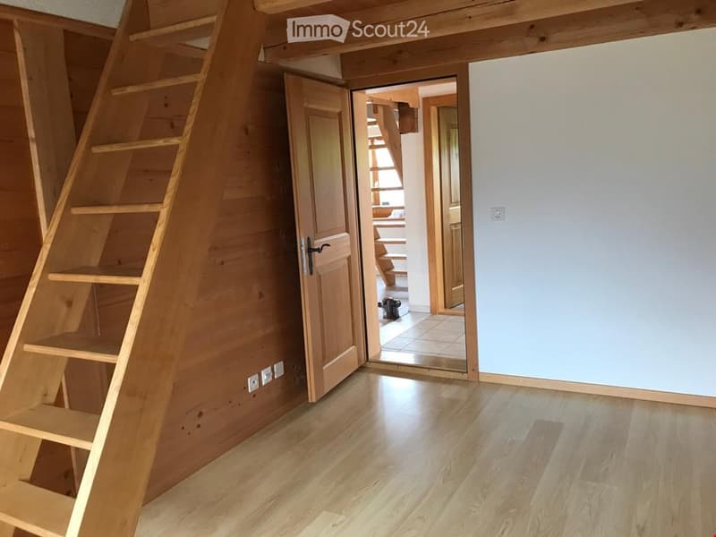 Empty room with wooden flooring, white walls, and a wooden staircase leading to another area. The room has a door that opens to a hallway with visible wooden stairs and a tiled floor.