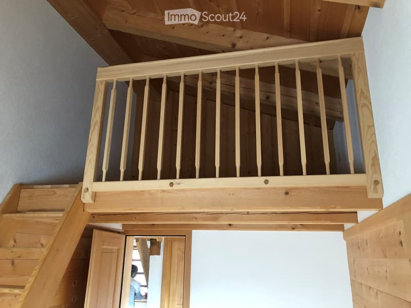 Wooden staircase with a wooden railing and open balusters, leading to an upper level with exposed wooden beams and a slanted ceiling.