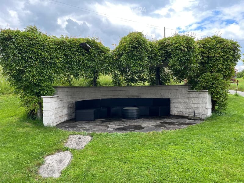 A small outdoor seating area with a stone wall enclosure, featuring a round table and benches surrounded by lush greenery and a grassy lawn.