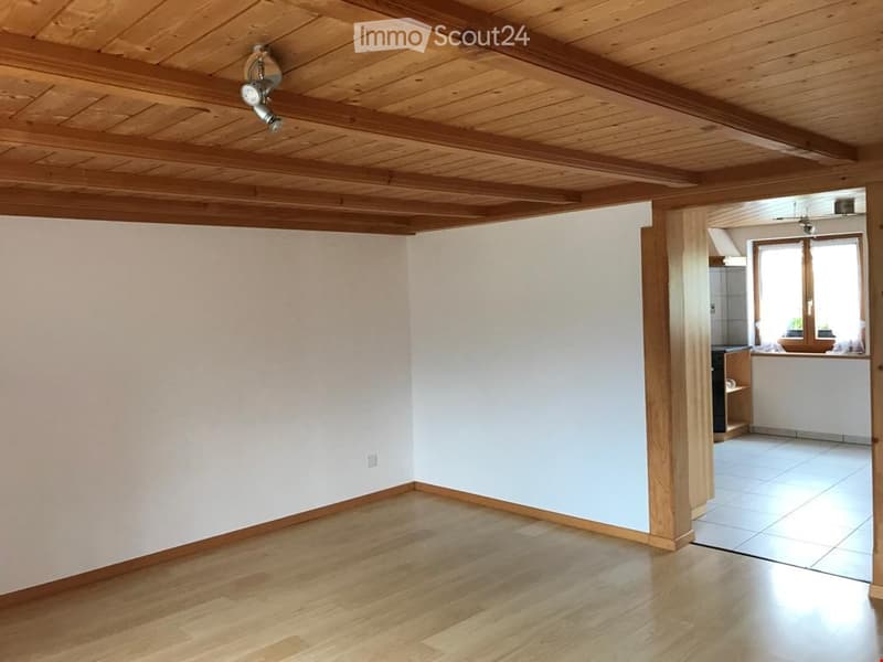 Empty room with wooden flooring and white walls, featuring a wooden ceiling with exposed beams and track lighting. An open doorway leads to a kitchen area with white cabinets and a tiled floor.