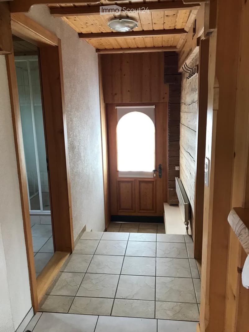 A narrow hallway with tiled flooring, wooden ceiling beams, and a wooden door at the end. The walls are painted in neutral tones, and there is a radiator on the right side.