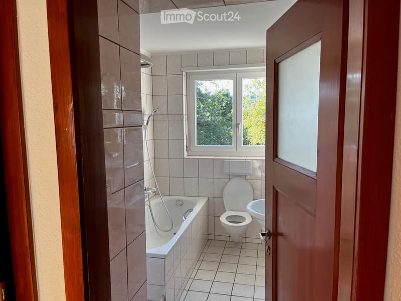 Bathroom with tiled walls and floor, featuring a bathtub, toilet, sink, and a window with a view of greenery.
