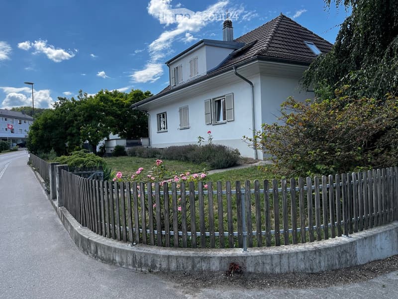 A two-story white house with a tiled roof, surrounded by a wooden fence and greenery. The house features multiple windows with shutters and a small garden area.