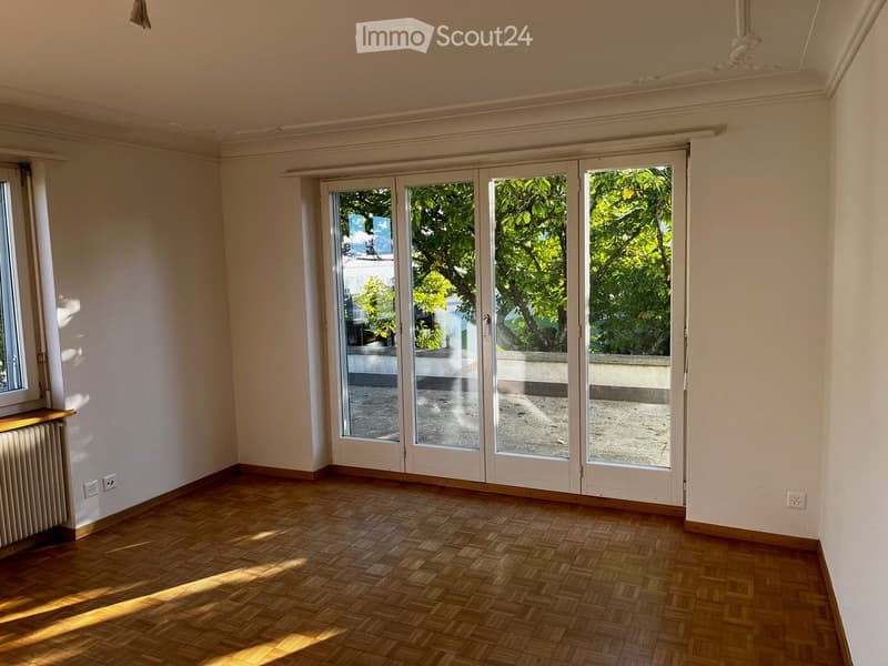 Empty room with wooden flooring, large sliding glass doors leading to a balcony, and a radiator on the left side.