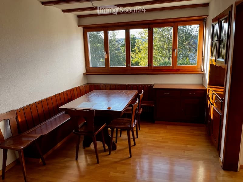 Wooden dining area with a long bench seating, a central table, and chairs, complemented by a wooden cabinet and large windows offering a view of greenery.