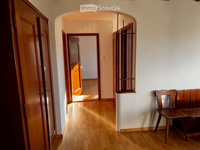 A well-lit hallway with wooden flooring, arched doorways, and a small wooden bench against the wall.