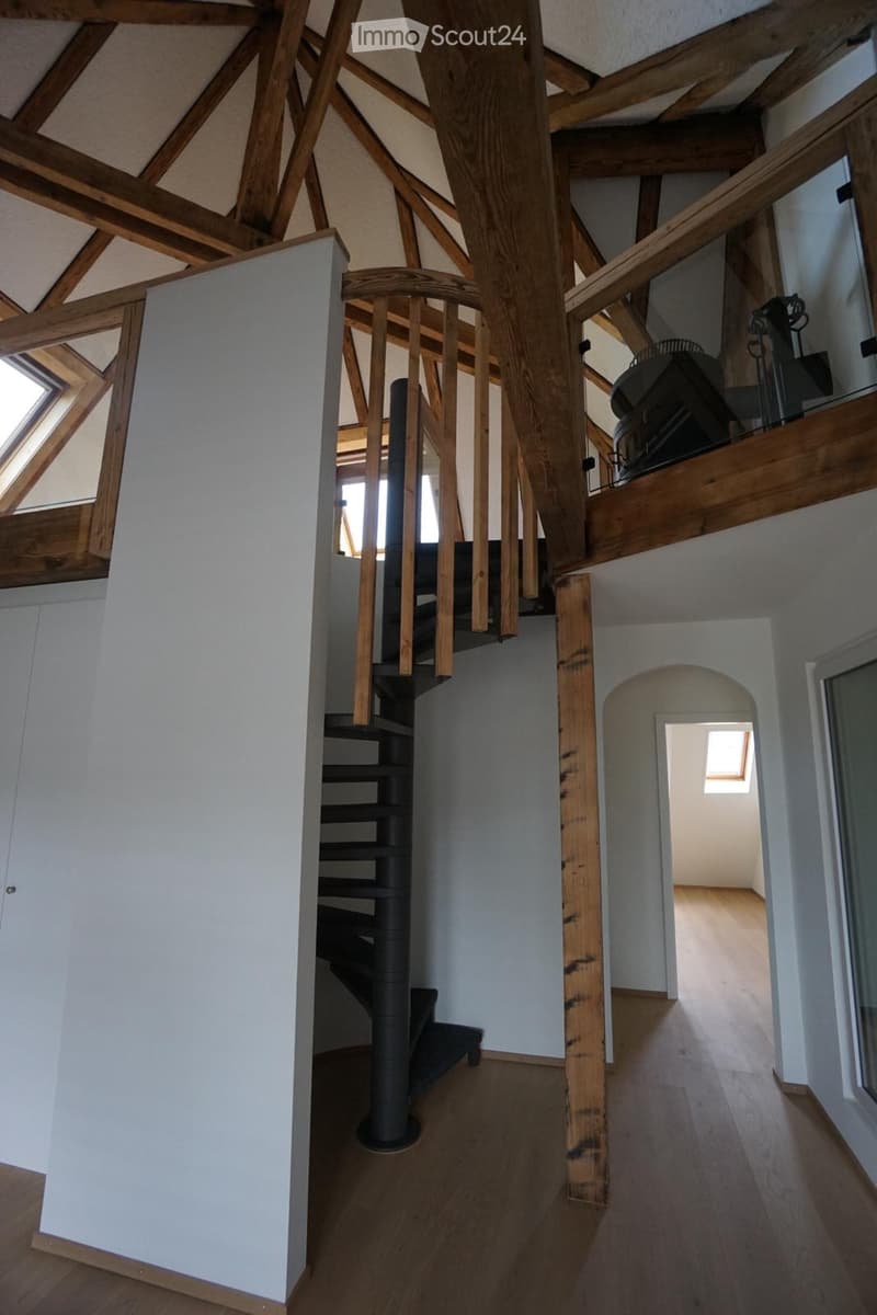 Spacious hallway with wooden beams, a modern spiral staircase, and exposed wooden beams on the ceiling. The floor is made of light wood, and the walls are painted in neutral tones. There is a small open area leading to another room with a skylight window.