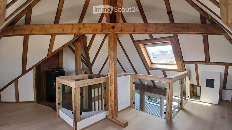 An attic space with exposed wooden beams, a skylight window, and a wooden staircase leading to a lower level. The room is empty, with no furniture or furnishings.