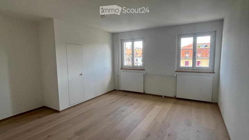 Empty room with wooden flooring, white walls, and two windows offering a view of neighboring buildings. Radiators are present along the wall.