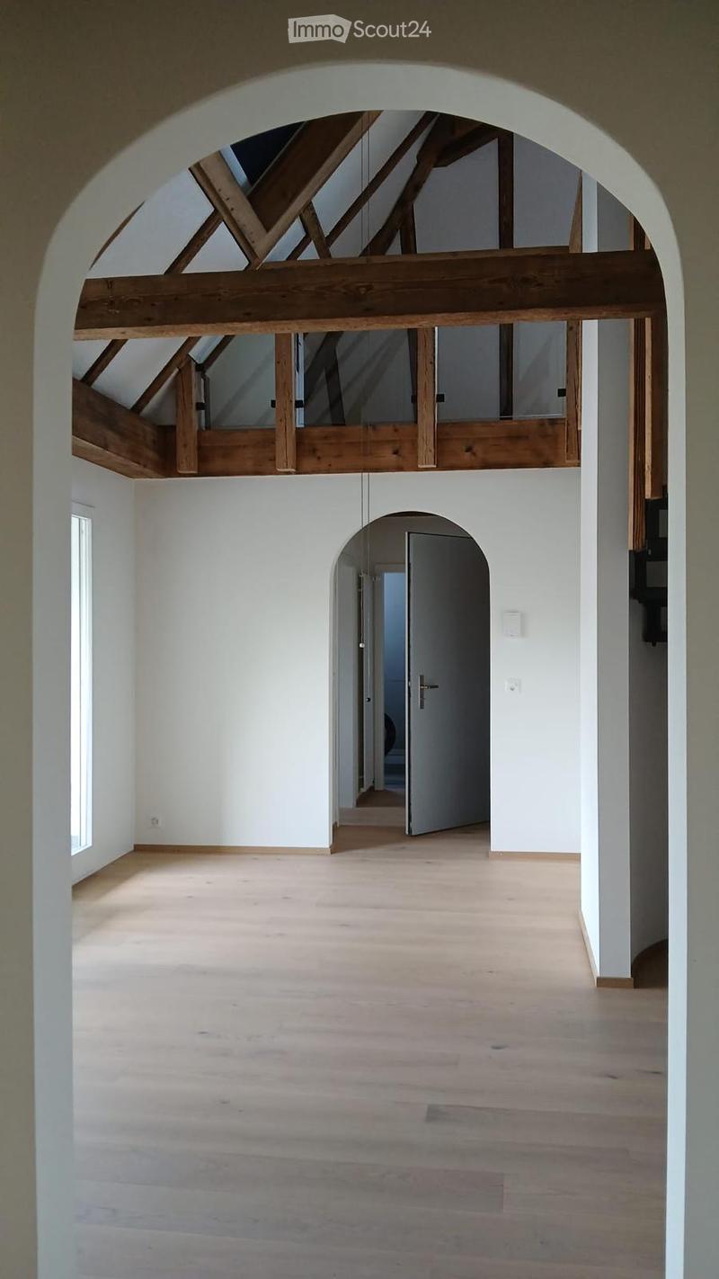 Empty room with wooden flooring, white walls, and exposed wooden beams in the ceiling. A doorway leads to another room.