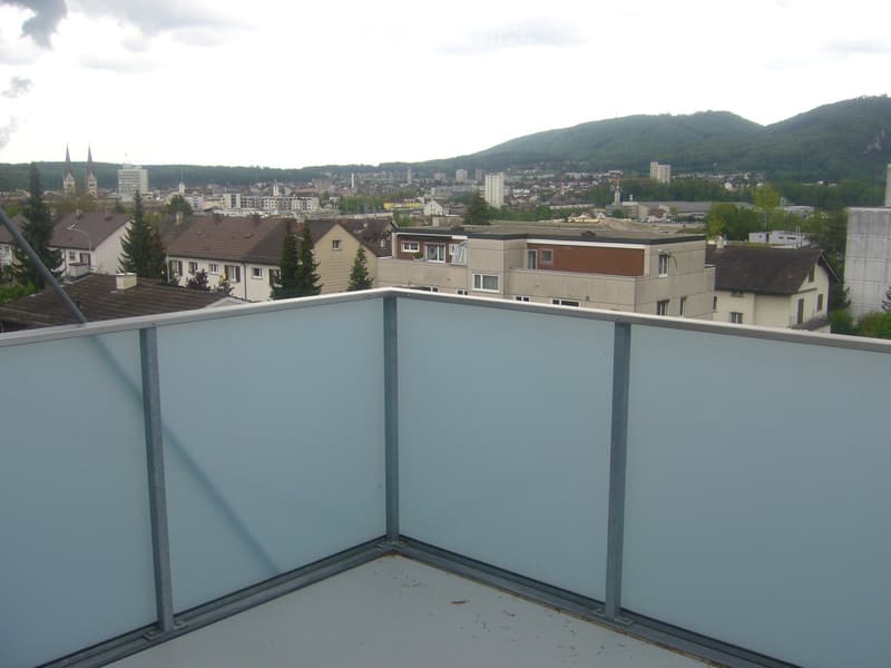 Roof terrace with low white walls, overlooking a cityscape with buildings, trees, and distant hills.