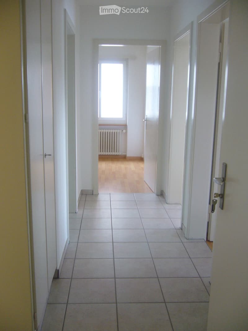 Empty hallway with white tiled floor and walls, featuring several doors and a window at the end.